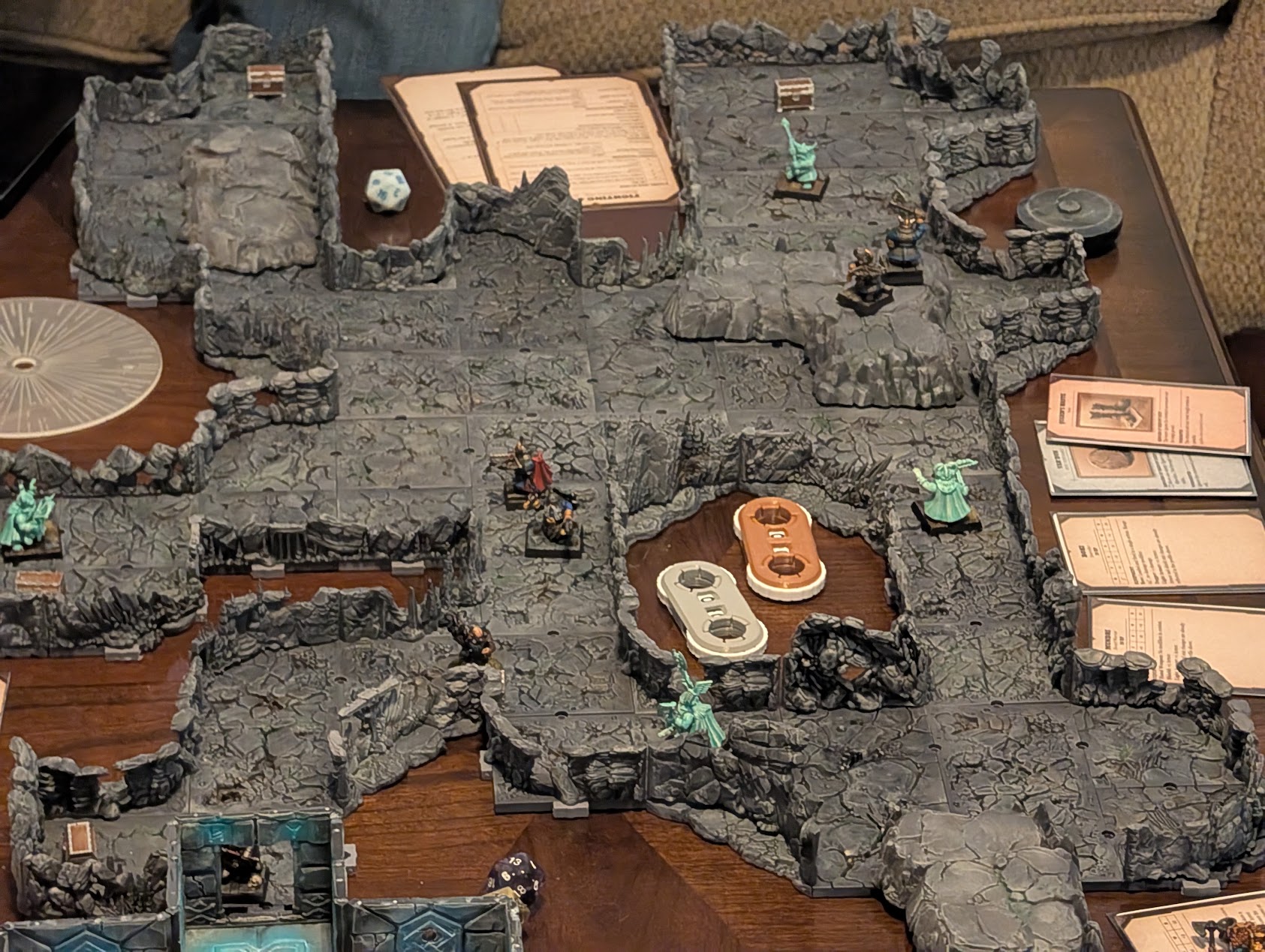 Overhead view of a winding cave layout with dwarf miniatures, pale green ghosts, and a furnished chamber.
